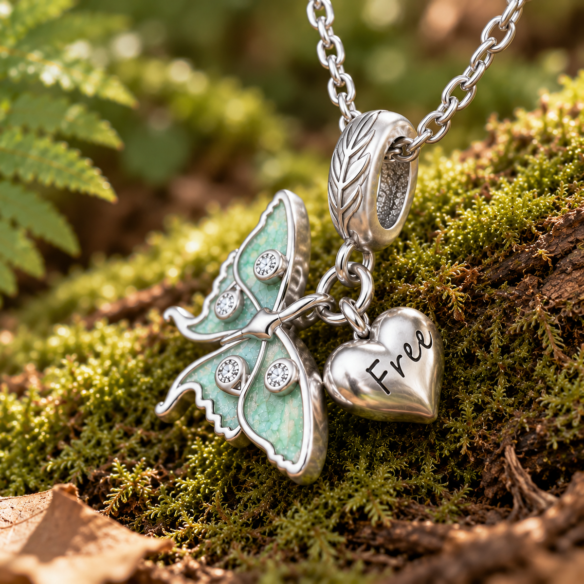 Silver moth and heart pendant necklace resting on mossy forest ground. The moth charm has pale mint-green enamel and small clear stones, and the heart charm is engraved with the word “Free.