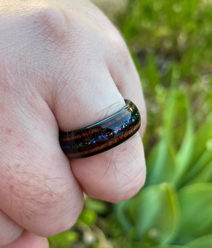 8mm black Tungsten supernova ring with Moon dust, crushed opal, holographic flakes, and acacia wood, worn on a man's hand in a natural setting.