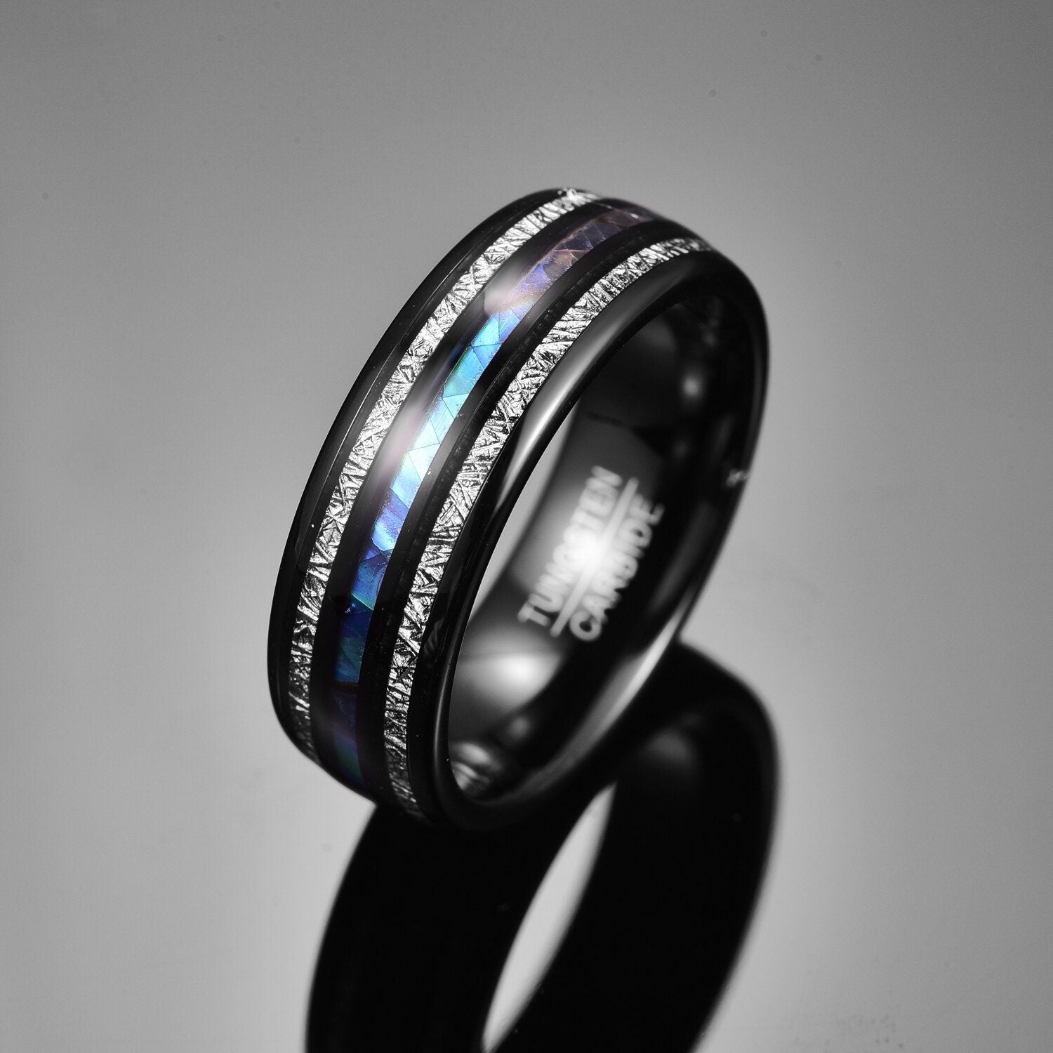 Black Tungsten ring with Gibeon meteorite and abalone shell inlays, displayed on a glossy black surface.