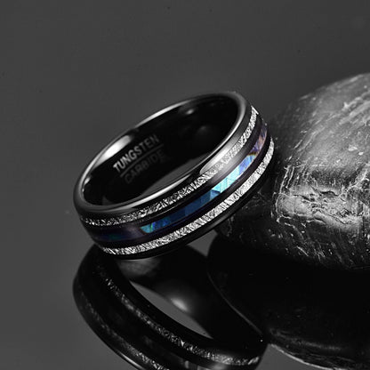 Black Tungsten ring with Gibeon meteorite and abalone shell inlays, resting on a reflective surface beside a textured stone.