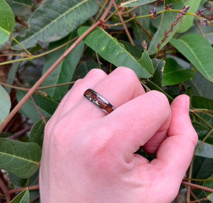 Hand wearing a koa wood band with silver Tungsten edges, surrounded by natural greenery.